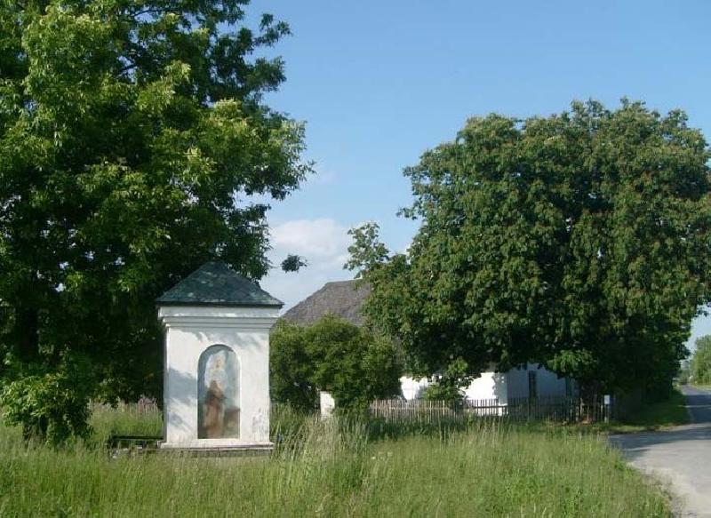 Biała kapliczka w Brodach Małych w trawie na tle drzew i błękitnego nieba